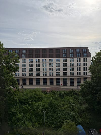 The image displays a daytime urban scene in Ingolstadt, Germany, featuring a modern building, a street, and abundant greenery under a bright, partially cloudy sky.
In the upper portion of the frame, the sky is bright and expansive, with diffuse sunlight originating from the upper left. A mix of wispy and more defined white clouds are scattered across the light blue-grey sky.
The central subject is a tall, multi-story building, likely an office or residential complex, with a distinct two-toned facade. The left side of the building features a darker, possibly brown or bronze-colored facade with numerous smaller, uniformly square windows. The right side presents a lighter-toned facade, appearing to be made of stone or concrete panels, characterized by larger, rectangular windows that reflect the sky and surrounding trees.
At the base of the building, a paved street or wide pathway stretches horizontally. A person on a bicycle, wearing dark clothing, is riding along this path in front of the building, heading towards the left side of the frame.
The lower and right-hand sections of the image are dominated by lush, dense green trees and foliage, providing a natural contrast to the urban architecture. Among the trees on the far right, a dark-colored car is partially visible, parked alongside a blue tarp or cover. A black lamppost stands amidst the greenery further to the right.
Notable details include a small red square sign with an indistinct white symbol visible on the lighter facade of the building, and another smaller white square sign with a black symbol, too blurry to read, near the ground level on the far right. The time of day is clearly daylight, indicated by the strong sunlight. The overall mood is calm and everyday.