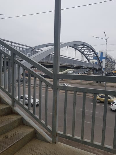 The image shows a pedestrian overpass with a metal staircase in the foreground. The overpass has a curved metal arch structure and is fenced with metal railings. Below the overpass, a multi-lane road is visible with several cars driving on it. In the background, there is a building with a large blue "H" logo and apartment balconies. The sky is overcast, suggesting a cloudy day. The location is identified as Бұлақты, Kazakhstan.