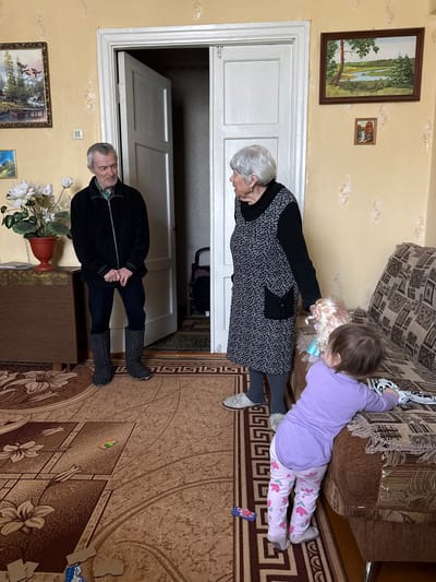 Location: Vyshegor, Russia

Scene: An interior view showing three people. An elderly man stands near a doorway, wearing a black jacket, dark pants, and felt boots. An elderly woman is leaning over a couch, wearing a grey dress and dark-colored sweater. A young child is lying on the couch, wearing a purple shirt and patterned leggings, playing with a doll. A brown patterned couch is behind the child and elderly woman. The room has a brown patterned carpet and beige walls. Paintings hang on the wall. A table with flowers is near the elderly man. A doorway is open behind the elderly man, revealing the interior of the next room.