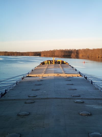 The image depicts the expansive deck of a barge or ship, likely transporting cargo, traversing a waterway. The deck is characterized by a gray, concrete-like surface dotted with circular hatches or access points arranged in parallel rows, receding into the distance. A low, blue railing runs along the edges of the deck. At the far end of the deck, there are some yellow structures and several people. The waterway is flanked by a forest on both sides. A red buoy is visible in the water to the right. The sky is clear with a gradient of light blue to a warmer, brighter hue closer to the horizon, suggesting either sunrise or sunset. According to the metadata, the picture was taken on the Volga river near Kurdiug, Russia.