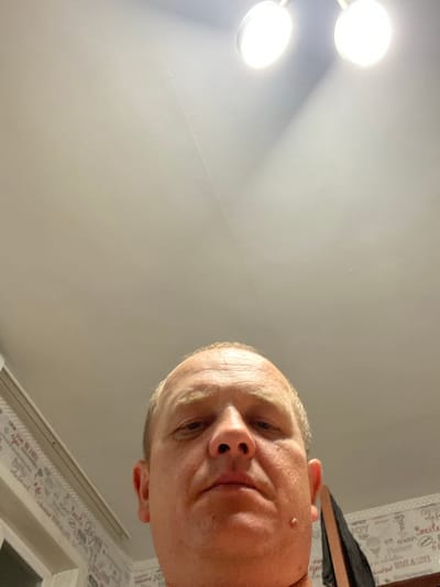 The media file is a close-up, low-angle selfie of a person's face, oriented sideways. The subject is an adult with fair skin and blond hair. Their head is tilted downwards towards the camera. One eye is visible, appearing to be looking slightly to the side. A small mole is visible on their cheek. The person's lips are slightly parted.
The background consists of a white ceiling, likely in an indoor setting, with two bright, circular light fixtures visible in the upper left corner. The lighting suggests it might be daytime or a well-lit interior.
There is a patterned wallpaper or fabric visible on the right side of the frame, featuring text and small graphics. Some of the visible text fragments include "Smile," "JOY," "LOVE," and "LIFE," suggesting inspirational or decorative phrases.
The overall impression is a casual, candid photograph taken from an unusual perspective, possibly of the person looking up towards a ceiling. No specific activity is discernible other than the act of taking a selfie. The setting appears to be a domestic or indoor environment.