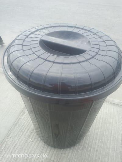This media file shows a black plastic trash can with a lid. The trash can is large and has a ribbed texture on its body and a patterned lid with a handle. The trash can is placed on a concrete surface, which appears to be outdoors. The lighting suggests it is daytime, and the weather is likely overcast or hazy, as there are no strong shadows. The background is blurred, but it seems to be an open area. There are no people clearly visible in the frame, though a small portion of a leg and shoe might be in the top left corner, suggesting someone might be near the trash can. The overall scene is mundane and focuses on the object itself. At the bottom of the image, there is visible text reading "TECNO SPARK 10c". The location context provided is Awoyaya, Nigeria.