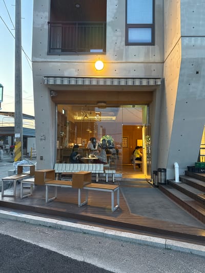 The image shows the exterior of a modern building, likely a cafe or small business, located in Toseong-myeon, South Korea. The building has a concrete facade with a large glass front. Inside, people are seated at tables. The building has a wooden deck area outside with chairs and tables. A utility pole is visible in the foreground. The sky is clear and the lighting suggests it is either early morning or late afternoon.