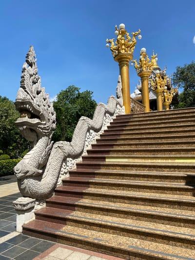 A view of a temple in Dĩ An, Vietnam presents a stone dragon-shaped statue that guards a staircase with granite stairs. Ornate gold lampposts with white globe lights appear adjacent to the stairs. Lush greenery and a clear blue sky form the backdrop.