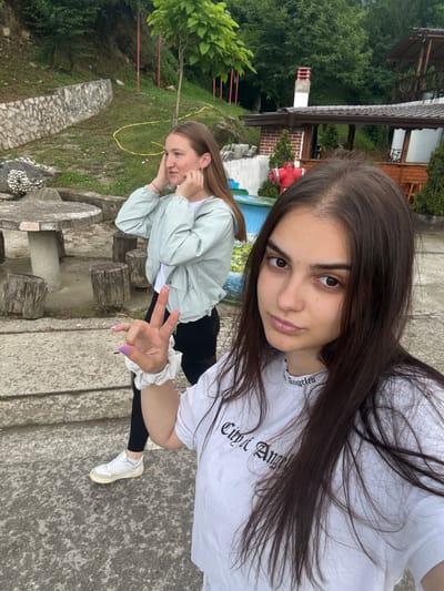 This media file captures two young women in an outdoor setting, likely a garden or yard, in Polena, Bulgaria. The image is oriented with the subjects and background rotated 90 degrees clockwise.
The primary subject, closer to the camera, has long, dark brown hair and is wearing a white t-shirt with the text "City Angeles" (or "City Angel") in black gothic font, black leggings, and white sneakers. She looks directly at the viewer with a slight smile and her right hand is raised, displaying a peace sign with light purple nail polish. A white scrunchie is visible on her wrist.
In the background, a second young woman with lighter brown hair is seen wearing a light green jacket over a white top. She has her hand near her face, possibly touching her ear, and her expression suggests a casual, perhaps contemplative, mood.
The scene is set on a concrete ground surface with a rustic outdoor seating area made of concrete and wooden stumps. Behind the second woman, a grassy slope with lush green foliage, trees, and bushes is visible, along with a yellow garden hose. In the far background, traditional-style buildings with brown roofs, a white chimney, and a brick wall section are discernible. A red object resembling a fire hydrant is also present near a building. The lighting indicates it is daytime, possibly with overcast skies, creating a soft, even illumination. The overall atmosphere appears relaxed and casual.