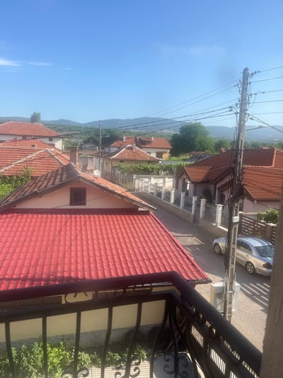 This is a vertical, eye-level, outdoor shot of a residential area in Krainitsi, Bulgaria on a clear, sunny day. The foreground is dominated by the bright red tiled roof of a house, with shadows cast by a dark metal railing in front of it.

Beyond this house, the scene opens up to reveal a street lined with other houses, many also featuring red tile roofs. A silver sedan is parked on the side of the road, with shadows from a nearby fence falling across it. A utility pole with numerous wires is a prominent feature in the middle ground, crossing the view from left to right. Green trees are scattered throughout the neighborhood, and in the distance, rolling hills under a bright blue sky suggest a rural or semi-rural setting.

The overall impression is one of a quiet, ordinary day in a European village. There is no visible text in the image. The lighting and clear sky indicate it is likely daytime, possibly late morning or early afternoon. The scene evokes a sense of peacefulness and everyday life.