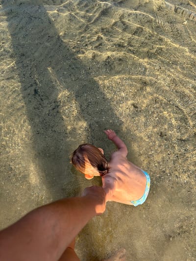 In Novalaise, France, a close-up, overhead shot captures a child lying in shallow, clear water, possibly at a lake or beach. The child, facing away from the camera and towards the left, has light brown hair and is wearing blue patterned swim trunks. Sunlight refracts through the water, creating dappled patterns on the sandy, pebbly bottom. The hand of an adult, presumably holding the child's arm, is visible in the foreground, also partially submerged in the water. The adult's arm is tanned and positioned to the left of the frame. The scene is tranquil, suggesting a moment of shared activity between an adult and child in a natural, watery environment.