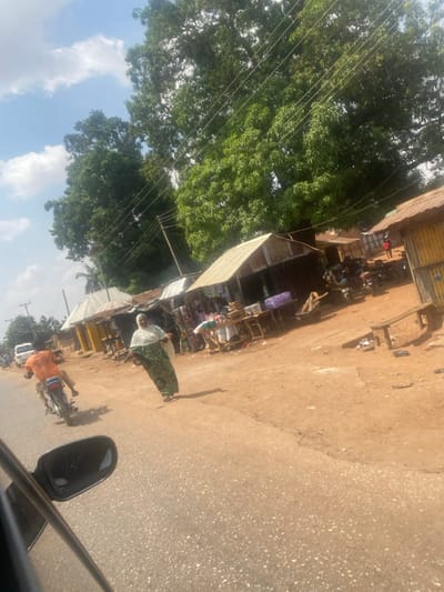 The image depicts a roadside scene in Tataura, Nigeria, under a bright, partly cloudy sky. A paved asphalt road occupies the lower left and central portions of the frame, with the black side mirror of a vehicle partially visible in the lower left foreground.
An adult female, wearing a long, patterned green garment and a white head covering, walks from right to left across the paved road. Behind her, on the left side of the road, an adult male rides a dark-colored motorcycle away from the viewer, dressed in an orange t-shirt and blue jeans. Further down the road on the left, a white vehicle is partially visible.
To the right of the paved road, a wide dirt shoulder features multiple makeshift market stalls or shops constructed with corrugated metal roofs and walls. Merchandise, including textiles and various packaged goods, is displayed on tables and hanging racks outside the stalls. Several motorcycles are parked near these structures. A wooden bench is positioned in front of the rightmost stall, which exhibits yellow/brown corrugated walls. In the background, beyond the stalls, indistinct figures are visible. Overhead utility lines are strung across the upper portion of the scene, above the stalls and large, dense green trees that dominate the upper frame.