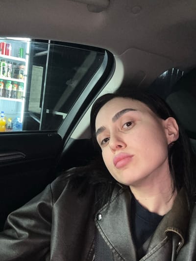 A woman in a black leather jacket sits in a car. She has dark hair, strong eyebrows, and pale skin. She pouts at the camera, sticking out her tongue slightly. Through the car window, the brightly lit interior of a store is visible, with shelves stocked with various food and beverage items. The car's interior is dark, and the overall scene has a casual, selfie-like quality.
