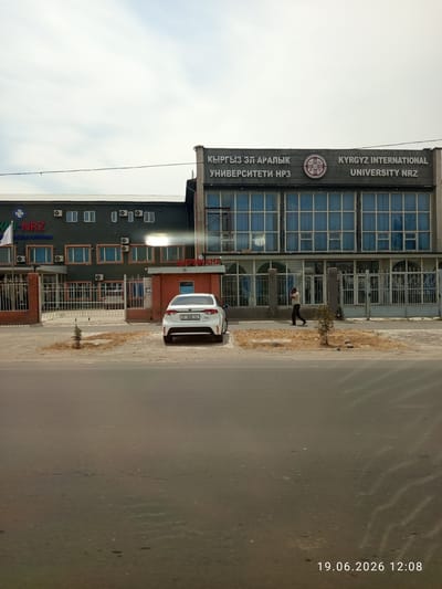 The image depicts a street scene in Novopavlovka, Kyrgyzstan, featuring the Kyrgyz International University NRZ. A white sedan is parked in front of the university entrance. A man is walking on the sidewalk in front of the university building. The university building is a two-story structure with large windows and signage indicating its name in both Russian and English. The surrounding environment appears to be a city street with a sidewalk and some vegetation. A date and time stamp at the bottom right corner of the image reads "19.06.2026 12:08".