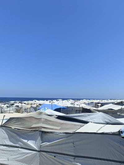 The image captures a sprawling refugee camp under a clear blue sky, likely during the daytime given the bright sunlight casting sharp shadows. The scene is dominated by rows upon rows of temporary shelters, predominantly white and gray canvas tents, constructed with poles and ropes. Some tents are pitched close together, creating a dense network of temporary housing, while others are more spread out. One prominent blue tent stands out amidst the neutral tones of the other shelters.
In the background, a thin line of the ocean is visible, suggesting the camp is situated near the coast. The water appears calm, reflecting the bright sky. The overall impression is one of a vast, makeshift community, likely established to house displaced people. The sheer number of tents and the simple construction materials evoke a sense of necessity and perhaps a precarious living situation. There are no visible people in the frame, but their presence is implied by the existence of the shelters. No text is discernible in the image. The setting appears to be a beach or sandy area, as indicated by the light-colored ground visible between the tents. The weather is fair and sunny, contributing to the starkness of the scene.