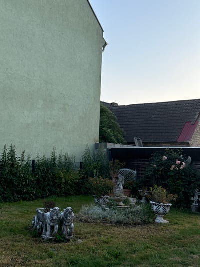 This is a full shot of a backyard in Wustermark, Germany. The scene is dominated by a large, pale green building on the left, its rough-textured facade filling a significant portion of the frame. To the right, a dark-tiled roof slopes down, with a section of red roofing visible on a smaller adjacent structure. A wooden fence runs horizontally across the middle ground, behind which are dense green bushes and a rose bush with pink blooms. In the foreground, on the grassy lawn, are several garden ornaments. Most prominent is a stone planter designed to look like a cart, pulled by two stylized lion-like figures. To the right of the cart planter is a small tiered fountain with a statue of a winged figure, surrounded by various smaller plants and pots, including a large white planter with black decorative markings. The sky above is clear and pale blue, suggesting it is either early morning or late evening.