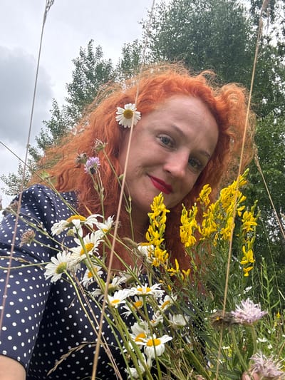 The image shows a woman with red, curly hair, her head tilted towards the camera, smiling and holding a bouquet of wild flowers. She is wearing a dark blue dress with white polka dots. She has red lipstick on. The bouquet consists of daisies, yellow broom flowers, and some other smaller purple and pink flowers. She is outdoors, with a background of green trees and a cloudy sky. The photographer is presumably close to the woman, as the perspective is from a low angle, looking up at her. There are no visible buildings or landmarks that would indicate a specific location within Privolzhsky, Russia, or any city for that matter. The scene appears to be a natural setting, possibly a field or meadow, on an overcast day.