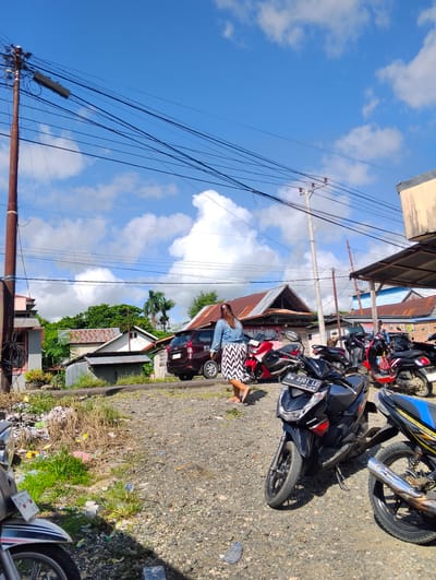 The image depicts an outdoor scene in the city of Pompanua, Indonesia, under a bright blue sky with scattered white clouds. The foreground is a gravelly ground with some patches of dry and green grass, as well as scattered debris. Several motorcycles are parked in the middle ground. A woman wearing a denim jacket and a patterned skirt is walking from right to left in the center of the image. Behind her, a dark-colored car is parked. The background shows several buildings with corrugated metal roofs, some overgrown vegetation, and utility poles with numerous wires crisscrossing overhead. The overall impression is of a somewhat cluttered and casually maintained urban or semi-urban environment.