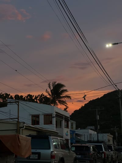 This image captures a street scene in San Juan del Sur, Nicaragua, during sunset. The sky displays a gradient of soft purples and oranges, with a few wispy clouds. Power lines crisscross the frame, a common sight in urban environments. A street lamp illuminates a portion of the sky on the right side.
In the foreground, several vehicles are parked along the street. The most prominent is a silver SUV with a license plate reading "M 132 209" and "NICARAGUA" below. To its right, other vehicles are partially visible.
Buildings line the street. On the left, a white building with orange-trimmed windows is visible. Adjacent to it is another building with a storefront, and further down, more structures can be seen. A palm tree with its fronds swaying in the breeze is positioned behind the white building.
A dark, tree-covered hill dominates the background on the right side of the image. The overall impression is a tranquil evening in a coastal town.