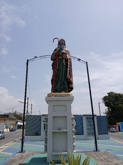 A colorful statue of Saint Joachim stands prominently on a tall, light-colored concrete pedestal in a public area of San Joaquín, Venezuela. The statue, oriented towards the viewer, depicts an elderly male figure with a long white beard and hair, dressed in a green tunic and a brown-patterned cloak. He holds a red shepherd's crook in his raised right hand and a golden, ornate object in his left. The pedestal features multiple rectangular recessed sections. In the immediate foreground, green and yellow spiky foliage grows from a tiled ground patterned with blue and green geometric shapes. Behind the statue, two tall black metal poles are connected by an arched crossbar. Further back, a light blue building with vertical corrugated panels displays a white informational sign, part of which reads "SAN JOAQUIN." A paved street extends across the midground, with a silver sedan parked on the left side and a white pickup truck on the right. Residential buildings and a vegetated hillside are visible in the background to the left, while trees and additional structures are present on the right. The scene is under bright daylight with a clear blue sky and scattered white clouds.