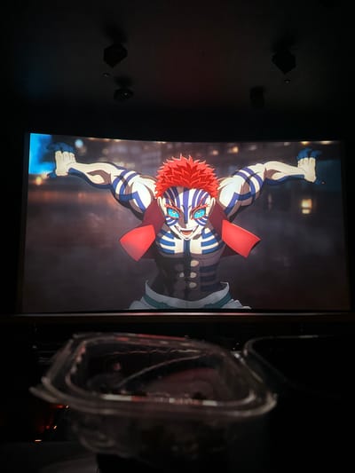 This image depicts a darkened movie theater in Paradise, United States, where a large, curved screen displays an animated character. The character, Akaza from *Demon Slayer*, is a muscular male with bright red spiky hair, prominent blue striped markings across his white skin, light blue eyes with white pupils, and sharp teeth. He wears a short red cape/shawl and a dark, armored midsection garment over white pants. His arms are outstretched to the sides, hands open, with blue glowing energy emanating from his left hand. His facial expression is aggressive, with an open mouth suggesting a roar. The background of the animated scene is a dark, blurred urban or industrial setting with faint distant lights. In the immediate foreground, a clear plastic clamshell-style container is partially visible, appearing to hold dark, granular contents, likely a snack. Above the screen, two black, cuboid speakers are mounted on the dark ceiling. The overall environment is dim, consistent with a cinema during a showing.