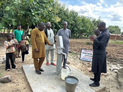 In the city of Afogo, Nigeria, a group of individuals, including several young children and adult men, are gathered around a newly installed water pump. The scene appears to be a communal area, possibly a village square or a designated spot for the water source, with a dirt ground and a few buildings in the background. The sky is partly cloudy.
The water pump itself is a hand-operated model, made of metal, mounted on a concrete base. Two buckets are placed on the base, one white and a bit smaller, the other a dark metal bucket. The white bucket contains clear water, while the metal bucket holds a larger quantity of water.
The people present are dressed in a mix of traditional and modern attire. A man in a bright yellow and black patterned robe stands closest to the pump, facing away from the camera. To his left, a man in a grey outfit with a similar buttoned collar is looking down, perhaps in prayer or contemplation. Further left, two young boys and a girl are looking towards the pump. Another man in a green t-shirt is standing behind them, observing.
To the right of the pump, a man in a black outfit, possibly a traditional robe with a split skirt, stands with his hands clasped, facing the pump. Another man, partially visible behind him, also appears to be dressed in dark clothing.
A white sign with black lettering is propped up on the ground behind the group on the right. The visible text reads "DONAT NORTHERN ASSOCIA WITH SUPPOR FOUNTAIN OF LIFE". This suggests the water pump may be a recent donation to the community.
The environment around the pump is characterized by dry, tilled earth, indicating it could be a farming area. Lush green trees and bushes are present in the background, providing a natural backdrop to the scene. The overall impression is one of a community gathering to inaugurate or inspect a new, vital resource.