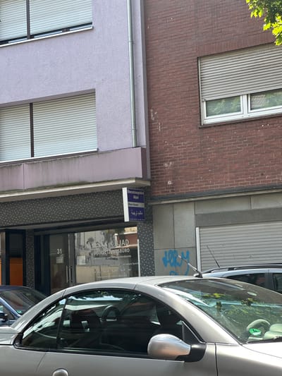The image shows a street scene in Hamm, Germany, on a sunny day. The perspective is from a car, looking towards a building. The building has a brick facade on the left and a lighter colored facade on the right, with several windows covered by rolling shutters.
On the right side of the building, there is a business with the name "BJAD ÜBERSETZUNGSBÜRO" visible on the glass door and a sign above it. The business offers translation and interpreting services in Arabic, English, and French, as indicated by the text on the door. There is also Arabic text on the sign and door. The building number "31" is clearly displayed on the glass.
A silver car is parked in front of the building, taking up a significant portion of the right side of the image. The car's windshield, side mirror, and part of the roof are visible. The reflection on the car's surface suggests a clear, bright day. Trees with green leaves are visible in the reflection on the windshield, indicating foliage nearby. The overall scene depicts an urban environment with commercial activity.