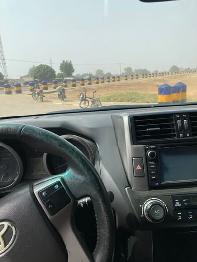 The image depicts a view from the driver's seat of a vehicle in Yan Dala, Nigeria. In the immediate foreground, the black steering wheel, partially obscuring the gauge cluster which displays 'D' for drive and '5916', is visible. To the right of the steering wheel, the vehicle's dashboard features air vents, an infotainment system, a prominent red triangular hazard light button, and climate controls including an 'A/C' button.

Beyond the windshield, a paved road surface is visible, alongside a reddish-brown dirt shoulder or median. A long line of cylindrical bollards, painted in alternating blue and yellow bands, extends into the hazy distance, marking a barrier or road division. Two motorcycles are parked on the paved area near these bollards, with a person standing adjacent to one of them. A black bicycle with a front basket is parked upright on the dirt area to the right of the bollards. In the background, under a pale, hazy sky, sparse trees, indistinct structures, and a large power line pylon on the far left are discernible.