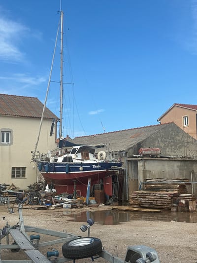 This image captures a dry-docked sailing yacht, identified as "Xenia," positioned within a boatyard or marine service area in Betina, Croatia. The yacht features a dark blue hull stripe, a white cabin superstructure, and a red antifouling bottom, and is elevated on multiple blue and metallic support stands. Its tall mast extends towards a clear blue sky with sparse white clouds. A rolled foresail is visible, and the stern displays a white lifebuoy and a small antenna. Several white fenders hang along the port side of the hull.
To the left of the yacht stands a two-story, light-colored building with a reddish-brown tiled roof and white-framed windows. Immediately to the right of the yacht, a single-story, grey, weathered building with a dark corrugated roof is present, which appears to be a workshop or storage facility. A red cylindrical tank is visible on its roof. Further right, a partial view of a second two-story building, light peach in color with a similar tiled roof and a white-framed window, is visible.
The ground is an unpaved surface of dirt and gravel, marked by several puddles of standing water. In the foreground, two grey metal boat trailers are present; one is partially obscured, while the other features a black spare tire with a silver rim resting horizontally on its frame. Scattered around the base of the yacht and the surrounding area are various items including stacks of raw lumber, several bicycles leaning against the left building, barrels (one brown, one blue), and general marine equipment or debris. No individuals are visible in the scene, which is brightly lit by natural daylight, indicating ongoing boat maintenance or storage operations.