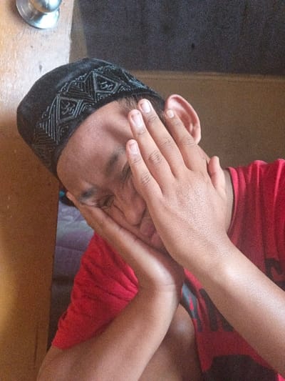An individual, identified as male, is positioned in an indoor setting within Dogsenang Gorphoog, Bhutan. The subject wears a black cap featuring intricate geometric patterns and a red t-shirt with a dark emblem partially visible on the right side of the chest. Both hands are raised, obscuring the face; the left hand covers the lower jaw and chin area, while the right hand covers the eyes and nose. Fingernails are visible and unpolished. The subject's head is tilted downwards and to their right. A portion of the left ear is exposed. The background is indistinct, consisting of a light brown vertical surface on the left, a darker, textured wall in the upper right quadrant, and a lighter brown wall segment. A metallic spherical object, possibly a doorknob, is situated on a light brown surface in the upper left corner. A purple-hued fabric is partially visible behind the subject's left arm.