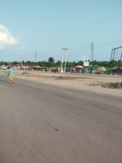 The image depicts a street scene in Uduagba, Nigeria. The foreground is dominated by a paved road with visible potholes and tire marks. On the left side of the road, a person in a blue shirt and patterned pants is walking across the street. The background reveals a bustling roadside market with numerous stalls, umbrellas, and palm trees. Several motorcycles are parked or in motion, suggesting activity. Power lines and utility poles stretch across the landscape, along with a tall transmission tower in the distance. The sky is clear blue with a few scattered clouds. The scene appears to be a typical commercial area with significant pedestrian and vehicular traffic.