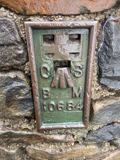 The image shows a benchmark in Beddgelert, United Kingdom. The benchmark is rectangular with a raised border, displaying a cross symbol on the left and the text "OS BM 3901" on the right. It appears to be made of metal, with a greenish-bronze color. The benchmark is embedded in a cobbled stone surface, with small pebbles and debris filling the gaps between the stones and the benchmark. The surface is wet, suggesting recent rain or moisture.