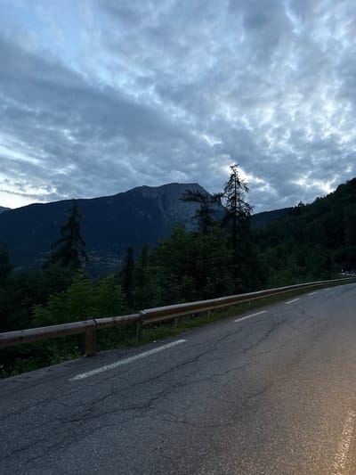 The image captures a winding asphalt road on the outskirts of Sainte-Foy-Tarentaise, France, under a cloudy twilight sky. A wooden guardrail runs along the left side of the road, separating it from a steep, forested mountainside. Tall evergreen trees and darker, denser foliage dominate the landscape, with a prominent, gnarled dead tree standing out against the sky. In the background, a large, dark mountain looms, its peak obscured by clouds. Power lines are visible stretched across the upper right portion of the frame. The road is marked with white dashed lines and shows signs of wear, with visible cracks and uneven patches. No people or vehicles are present in the image. The overall scene suggests a remote, natural setting at dusk.