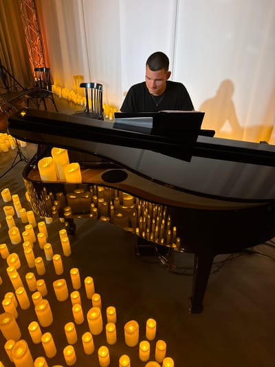 A young man with short dark hair is playing a grand piano. He is wearing a black t-shirt and a silver necklace with a cross pendant. The piano is black and appears to be a concert grand. The scene is illuminated by numerous flickering LED candles that surround the piano and are placed on the piano's surface. The background consists of draped curtains in warm tones and a white wall where a shadow of the man is cast. Two dark wooden chairs with metal frames are visible to the left of the piano. The floor is dark. The image is taken from a slightly elevated perspective, looking down at the scene.
