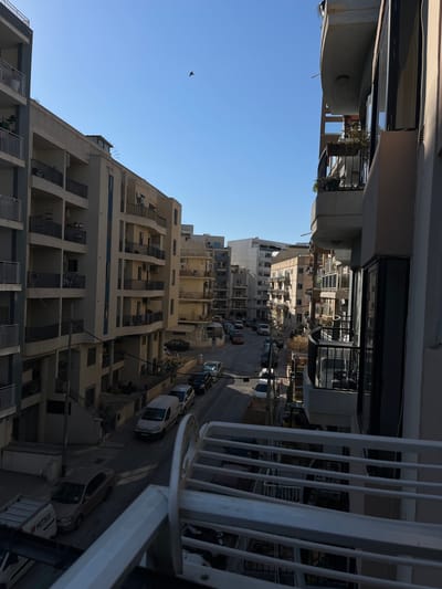 A vertical, medium shot captures a street scene in a city under a clear blue sky. The perspective is from a balcony, looking down and across at buildings lining a street. The buildings are multi-story apartment complexes with balconies and awnings, varying in color from light beige to yellow. A few cars are parked on the street, including a white van, a silver hatchback, and a dark sedan. The street is paved and appears to be on a slight incline. There is a lone bird in flight in the upper portion of the sky. The scene is illuminated by bright sunlight, casting shadows on the street and buildings.