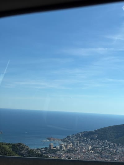 This image captures an elevated, panoramic view of the city of Budva, located in Montenegro. The scene is dominated by the vast expanse of the Adriatic Sea, stretching out to meet a clear blue sky dotted with faint wisps of clouds. In the foreground and mid-ground, the city of Budva unfolds along the coastline, characterized by a dense arrangement of buildings with terracotta rooftops and modern high-rises. A harbor filled with numerous boats and yachts is visible, along with a prominent peninsula featuring historical architecture. The cityscape is nestled against a backdrop of lush, green, mountainous terrain. The perspective suggests the photo was taken from a high vantage point, possibly from the surrounding hills or a higher elevation within the city.