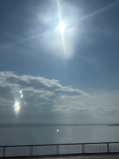 The image depicts a wide body of water under a bright, cloudy sky. The sun is visible as a strong, lens-flared light source in the upper center of the frame, casting bright rays and lens flares. Wispy white clouds are scattered across the sky, some appearing denser and more defined. In the lower portion of the image, there is a dark railing with vertical bars, suggesting a balcony, bridge, or elevated viewpoint. Beyond the railing, the water stretches to a distant horizon, where a faint landmass is visible. The overall scene is a landscape with natural elements and a man-made structure in the foreground. This image does not contain any people or specific actions. The location is not identifiable from the visual information provided in the image.