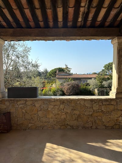 The image presents an outdoor scene captured from within a covered veranda or loggia in Mougins, France. The foreground features a light-toned, possibly tiled, floor partially illuminated by sunlight, casting distinct shadows. A robust, dry-stacked stone wall, composed of irregular light brown and tan stones, extends horizontally across the lower portion of the frame. A dark grey, rectangular planter box rests centrally atop this stone wall. The overhead structure includes a large, unfinished wooden beam spanning the width of the view, supporting a ceiling of exposed reddish-brown clay barrel tiles set between dark wooden rafters. Light-colored stone columns flank the left and right edges of the frame, delineating the viewing area. A dark brown fabric bag is partially visible on the floor in the bottom-left corner.

Beyond the covered area, a clear, bright blue sky dominates the upper background. The mid-ground reveals a sunlit, undulating landscape rich with diverse vegetation, including a prominent olive tree with silvery foliage on the left. Various other trees and shrubs, some with reddish-brown leaves and others evergreen, fill the scene, punctuated by patches of vibrant red flowering plants. In the distance, a traditional house with light-colored stone walls and a terracotta tiled roof is visible, partially obscured by greenery. To its right, a small, rustic stone shed with an arched wooden door is nestled amongst the trees. Further elements of distant architecture with tiled roofs and subtle mountainous contours are discernible on the horizon. No individuals or dynamic actions are depicted.