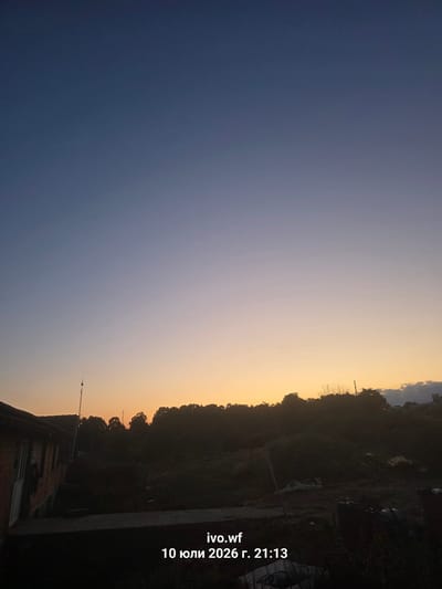 The image captures a twilight scene at dusk, likely in the rural outskirts of Chal, Bulgaria. The sky transitions from a deep blue at the top to a soft gradient of pale orange and yellow near the horizon. A faint star is visible in the upper portion of the sky.
In the foreground, a dark silhouette of trees and vegetation obscures much of the ground. To the left, a brick building with a white door is partially visible, suggesting a residential structure or outbuilding. A tall, thin pole with some wires extends upwards from near the building. The overall impression is one of a quiet, possibly undeveloped area as nightfall approaches.
The text overlay in the bottom center of the image reads "ivo.wf 10 юли 2026 г. 21:13", indicating the date and time the photograph was taken, July 10, 2026, at 9:13 PM. The Cyrillic script suggests a location where the Bulgarian language is used.