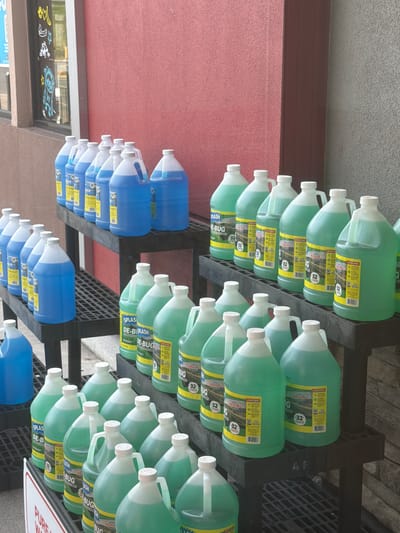 The image shows a retail display of windshield washer fluid and bug remover fluid jugs. The jugs are arranged on black plastic shelving units outdoors, in front of a building with a reddish-pink textured wall and a gray textured wall. The display is located at the Circle K convenience store in Raphine, Virginia. The jugs vary in color from blue to green and are of different sizes, mostly gallon jugs. Some of the labels are visible, showing brands like "Splash" and "Bug Repeller." The scene appears to be a sunny day. There are no people visible in the image.