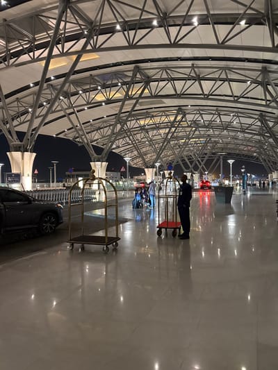 An indoor scene showcases the ceiling's architectural steel structure with repetitive triangular patterns. A man in dark attire pushes a cart through a shiny, reflective floor, alongside a line of empty luggage carts. Beyond the immediate interior, lights and outlines of buildings are visible against a dark sky. A black vehicle with its tire visible rests near a set of luggage carts.