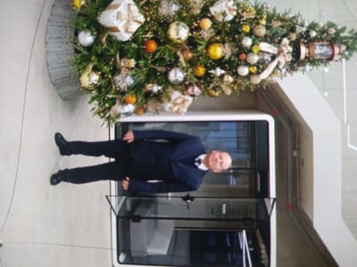 A man in a dark suit and tie stands in front of an open doorway, partially obscured by a Christmas tree adorned with ornaments and gift boxes. The tree, positioned to the left of the frame, is situated in a grey, woven planter. The man's legs are visible, and he appears to be standing with his hands at his sides. The doorway leads into a modern structure with glass panels. The surrounding environment appears to be an indoor public space with light-colored flooring. The image is oriented vertically, with the scene rotated 90 degrees clockwise from a standard horizontal perspective. The location is Birlik, Uzbekistan.