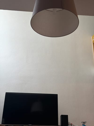 This image depicts an interior room in Eschau, France. The scene is dominated by a light-colored, textured wall (likely off-white or light grey) occupying most of the frame. Suspended from the white ceiling, in the upper portion of the image, is a cylindrical fabric lampshade, beige or light grey in color, with its interior visible. Below, a large, rectangular television with a dark, reflective screen is centered at the bottom of the frame. To the immediate right of the television, a black, rectangular electronic device (possibly a speaker or gaming console) stands upright. Further to the right, a small, blue and silver portable burner or stove is visible, positioned next to some crumpled white material. No individuals, actions, or direct interactions are visible within the static scene.