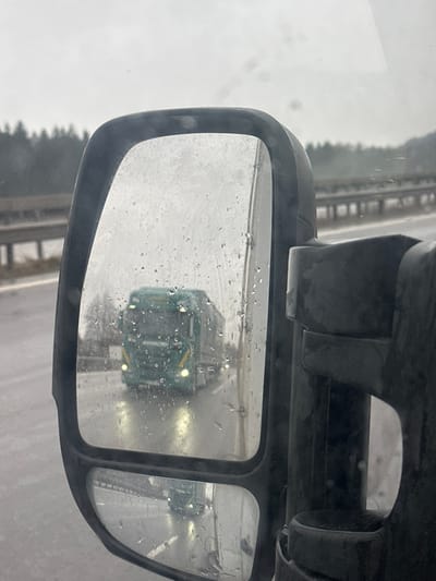 The image depicts a rainy daytime scene on a highway in Irschenberg, Germany, viewed from the interior of a vehicle looking towards its passenger-side mirror. The mirror, covered with numerous water droplets, reflects a large, green semi-truck with illuminated headlights and yellow accent stripes, traveling on a wet multi-lane asphalt highway. The truck's lights create bright reflections on the road surface. In the background, beyond the highway, a metal guardrail is visible, followed by a dense line of coniferous trees under an overcast grey sky. The dark frame of the vehicle's mirror assembly is prominent on the right side of the image. No individuals are directly visible.
