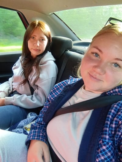 Two young women are seated in the back seat of a car, likely traveling through the city of Bogorodsk, Russia, as indicated by the presence of a Russian license plate visible on the car. The woman in the foreground, on the right, has fair skin, blue eyes, and blonde hair. She is wearing glasses on her head and a light-colored shirt under a blue and pink plaid jacket. She is smiling at the camera. The woman in the background, on the left, has light brown hair and brown eyes. She is wearing a light gray hooded jacket and is looking at the camera with a neutral expression. Both women are wearing seatbelts. The interior of the car is visible, including the headrests and a portion of the back window, which shows a blurry green, outdoor landscape, suggesting they are on a road with trees. A blue and white patterned blanket or scarf is visible between the two women. The lighting suggests it is daytime, possibly late afternoon due to the warm tones in the image.