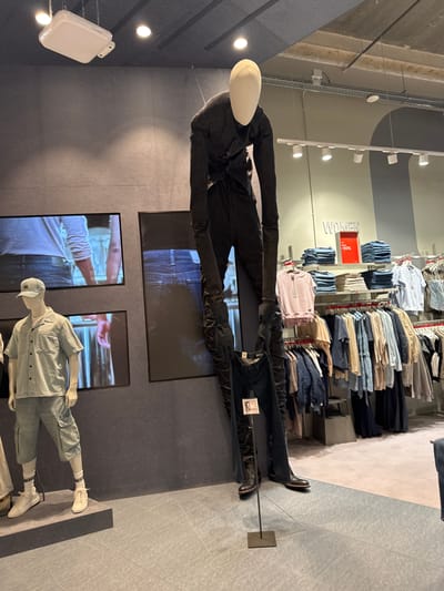 The image depicts a clothing store, likely H&M based on the "WOMEN" signage and red sale sign, located in The Leidschendam Mall, Netherlands. In the foreground, a tall, slender mannequin is posed wearing dark-colored clothing, holding what appears to be a pair of dark jeans. To the left, another mannequin is displayed wearing a light-colored short-sleeved shirt and shorts, a cap, and white sneakers. Behind these mannequins, screens display images of people wearing denim. The background shows racks of clothing for women, including stacks of folded jeans and shirts, and hanging garments. The store environment is well-lit, with overhead lighting visible. The floor is a light grey color with some scuff marks.
