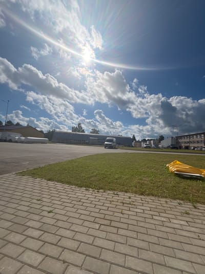 The image shows a bright, sunny day in Sauleskalns, Latvia. The sun is shining intensely, creating a strong lens flare with visible rays of light extending across the blue sky, which is dotted with fluffy white clouds. The scene is oriented vertically, with the sky dominating the left half of the frame.
On the right side of the image, a paved pathway made of grey interlocking bricks runs parallel to a strip of green grass. Beyond the grass, there are several buildings visible. One building appears to be a long, industrial-style structure with rows of windows. Further down the path, a few vehicles, including vans and what looks like a truck, are parked. In the foreground, to the right of the grass, a large object covered in yellow material is partially visible. The overall setting appears to be an outdoor area, possibly a park, an industrial estate, or a commercial zone.
The lighting suggests it is daytime, likely late morning or afternoon, given the sun's angle and brightness. The weather is clear and sunny, with a few clouds adding texture to the sky. There are no people visible in the frame, and the focus is on the landscape and architecture. No text is discernible in the image.