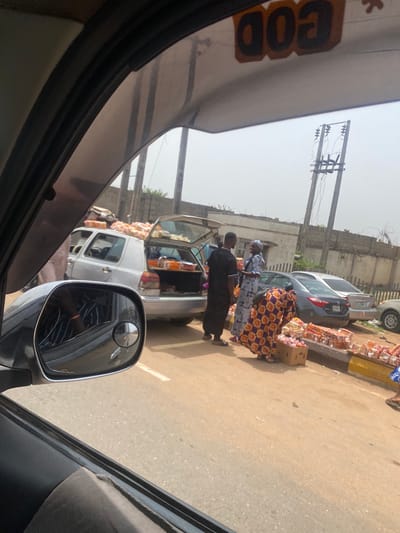 Captured from inside a vehicle, the image shows a street scene in Alakia, Nigeria. A white commercial vehicle with its trunk open is parked alongside a road filled with various goods. Several people, including at least two women in patterned clothing, are interacting with the merchandise. A grey car is also parked near the vendor's space. The environment appears to be a bustling street with buildings and utility poles visible in the background. The overall setting suggests a typical roadside market scene in Alakia.