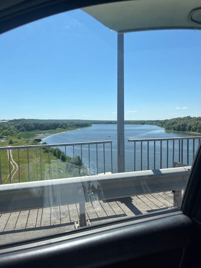 The image captures a scenic view from what appears to be a high vantage point, possibly a bridge or tower, overlooking a wide river or lake. The scene is bathed in bright daylight under a clear blue sky with a few wispy clouds, suggesting fair weather, likely mid-morning or early afternoon.
The foreground is dominated by the structural elements of the viewpoint, including a metal railing and a large support beam, which partially obstruct the view. The railing is made of vertical bars with horizontal supports, creating a repeating pattern. To the right, a dark, curved surface, possibly the interior of a vehicle or structure, frames the view.
Beyond the railing, the main subject is a vast body of water, reflecting the blue sky. Along the far bank of the water, a dense, green treeline stretches as far as the eye can see, indicating a heavily forested area. A narrow strip of land or a sandbar is visible in the water, also covered in vegetation. On the closer bank, there are patches of green grass and some distant trees.
Several small dark shapes on the water could be boats or people, though they are too small to be definitively identified. On the grassy area of the closer bank, there are faint outlines of paths or tracks. The overall impression is one of a serene natural landscape viewed from an elevated man-made structure. There is no visible text in the image. The location context provided, Yadrin, Russia, suggests this is a view of a Russian waterway, possibly the Volga River or one of its tributaries.