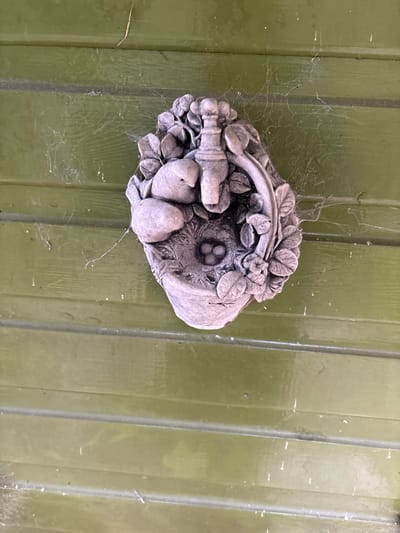 The image displays a weathered, grey, decorative wall ornament mounted on horizontal green wooden paneling in Stiperstones, United Kingdom. The ornament depicts a naturalistic scene, featuring two small, sculpted birds positioned near a nest containing three distinct eggs. Above the birds and nest, a stylized spigot or tap is centrally placed, flanked by floral and leaf motifs that extend around the perimeter, suggesting a basket or planter design. The entire scene is static, with no discernible actions or interactions involving animate subjects. The green wooden paneling exhibits signs of age or exposure, including faded paint and dust accumulation. Multiple delicate spiderwebs are stretched across the green surface, particularly visible to the left and above the ornament, indicating an outdoor or semi-sheltered environment. Small particles of debris are caught within these webs and on the wooden surface.