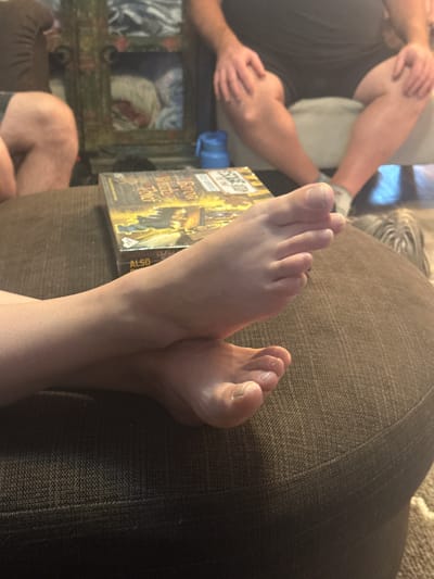 The image captures a close-up view of two bare feet resting on a textured brown surface, possibly a couch or ottoman. The feet are crossed at the ankles, with one foot positioned slightly above the other. The skin appears smooth, and the toenails are visible. In the background, a board game box is partially visible, along with parts of people's legs and torsos, suggesting a casual indoor setting. There is a blue object, possibly a water bottle, and a blurry glimpse of a dog in the far background. The overall scene implies a relaxed atmosphere, with people engaged in leisure activities at home. The image is not specific to any location in the Town of Cochrane, Canada, or any other city.