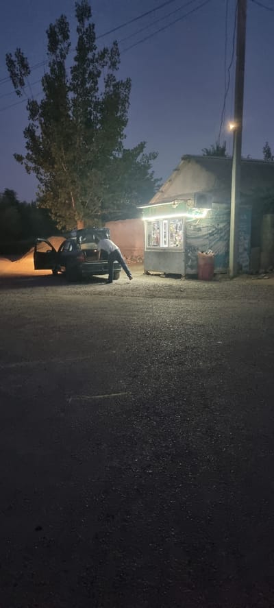 The image depicts a dimly lit scene in what appears to be a rural or semi-urban area of Khovaskor, Tajikistan, likely during dusk or early night. The primary subject is a person, dressed in a white shirt and dark pants, bending over into the open trunk of a dark-colored car. The car's trunk is illuminated by an internal light, casting a warm glow on the pavement. The car's rear passenger door is also open.
Adjacent to the car is a small, illuminated storefront or kiosk. It features glass display windows showcasing various products, and a bright, cool-toned light illuminates the front of the establishment. A large, reddish-orange bag is visible to the right of the storefront. Behind the storefront, a portion of a building is visible, with a light pole and overhead wires stretching across the frame. To the left of the storefront and car, a dense tree with dark foliage stands.
The ground is paved with asphalt, showing some texture and slight reflectivity, suggesting it might be damp. The overall atmosphere is subdued, with the artificial lights from the storefront and car providing the main sources of illumination against the darkening sky, which is a deep blue. No discernible emotions are conveyed by the person, as their face is obscured. The setting suggests a moment of interaction with the car or unloading/loading of items from the trunk, possibly in a late-night convenience store context given the storefront. The time of day is clearly evening or night.