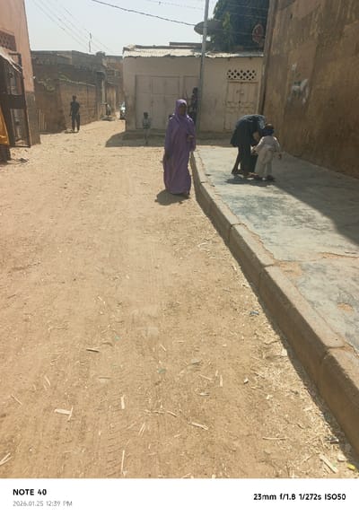 The image depicts a daytime street scene in Tudun Wazurchi, Nigeria. The foreground and midground are dominated by a dry, light brown dirt road with visible tire tracks and scattered wood chips and debris. A raised concrete sidewalk with a pronounced curb runs along the right side of the frame, showing signs of wear and minor cracking.

Multiple individuals are present. In the midground, right of center, a person wearing a long, flowing purple robe and head covering walks from left to right, casting a shadow to their right. Behind and to the right of this person, closer to the sidewalk curb, another individual in dark clothing is bent over, facing right, appearing to interact with a small child. The child, dressed in light-colored clothing and wearing blue goggles or eye protection, stands upright, facing right. Further down the dirt road on the left, a person in dark clothing walks away from the camera. Another small child, dressed in a light green top and white pants, is visible further down the road near a white building.

Buildings line both sides of the street. On the left, there is a light-colored building with an apparent open doorway or recessed entrance, followed by a low wall and more residential structures. On the right, a tall, weathered ochre-colored wall extends into the background, casting a prominent shadow across the sidewalk. In the midground, a white building with a corrugated metal roof and two large, light-colored double doors is visible, with another light-colored building featuring a patterned ventilation block and a door to its right. Electrical wires are strung across the street, and a satellite dish is mounted on a pole near the white building. The sky is clear and bright, indicating direct sunlight at what the embedded metadata identifies as 12:39 PM.