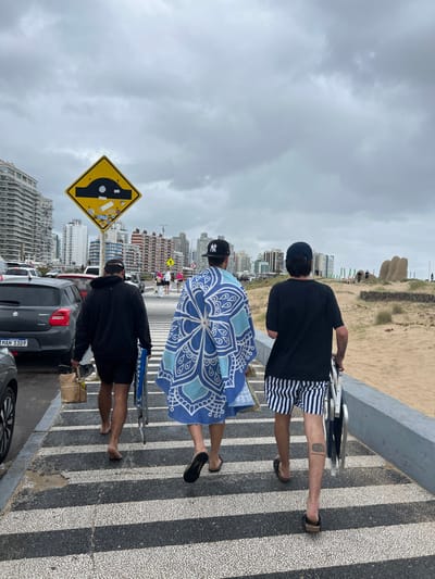 The image captures three individuals walking along a striped paved pathway next to a sandy beach under an overcast sky in Punta del Este, Uruguay. The scene appears to be taken during the daytime, with muted light due to the clouds.
In the foreground, a prominent yellow, diamond-shaped road sign indicating a humped bridge or speed bump is visible, covered with several small stickers. Directly behind it, a dark-colored Suzuki Swift car is parked, displaying the license plate "MAN 1309".
The three subjects are walking away from the camera. The closest person wears a black hoodie, black shorts, and is barefoot, carrying a box or bag. The next individual wears a black baseball cap with an "NY" logo, a dark top, and black sandals, and is holding a large, round beach towel or umbrella featuring an intricate blue and white mandala-like pattern. The third person, furthest from the camera, also wears a black "NY" baseball cap, a black t-shirt, striped swim shorts, and flip-flops, carrying folded beach chairs. A tattoo is visible on this person's right calf.
The walkway has a distinctive pattern of alternating black and white stripes. Adjacent to the path is a wide sandy beach. In the distance, a well-known sculpture of giant fingers emerging from the sand, commonly referred to as "La Mano," is clearly visible on the beach. Beyond the beach and sculpture, a dense urban skyline of tall buildings stretches along the coast. The activity suggests people are heading to or from the beach, possibly for relaxation or leisure, despite the cloudy weather.